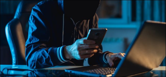 Um criminoso envolto em sombras usando um smartphone para fazer uma ligação na frente de um laptop.