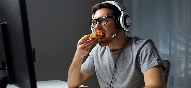 Um homem sentado na frente de um computador, usando um fone de ouvido e comendo pizza.
