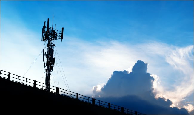 Uma torre de celular 5G contra um céu azul com nuvens brancas. 