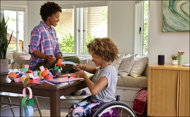 Uma criança em uma cadeira de rodas fazendo artesanato com a mãe em uma mesa com um Amazon Echo nas proximidades.