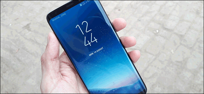 Uma mão segurando um Samsung Galaxy S8 com tela sensível ao toque mostrando a hora e a data.