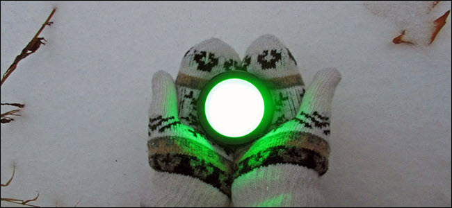Um par de mãos enluvadas segurando um botão Echo brilhando em verde sobre a neve.