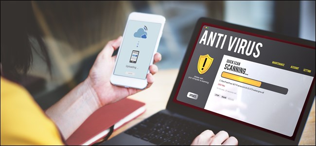 Uma mão segurando um smartphone que executa um antivírus, ao lado de um laptop em uma mesa que também executa uma verificação antivírus.