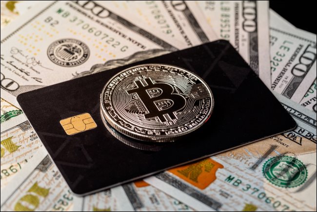Um token físico de Bitcoin em cima de um cartão de crédito sobre notas de $ 100.