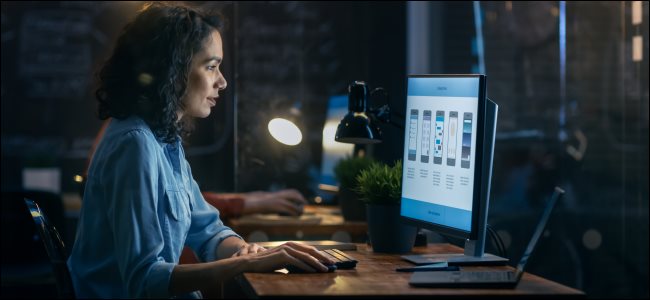 Uma mulher trabalhando em um computador desktop, com um laptop aberto ao lado dele. 