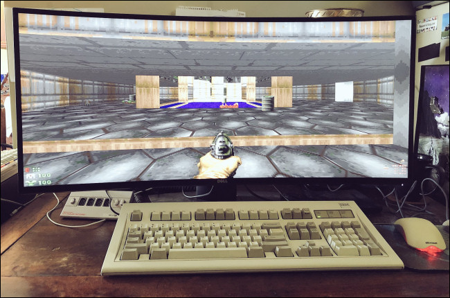 "Doom" em um monitor widescreen atrás de um teclado e mouse de mesa. 
