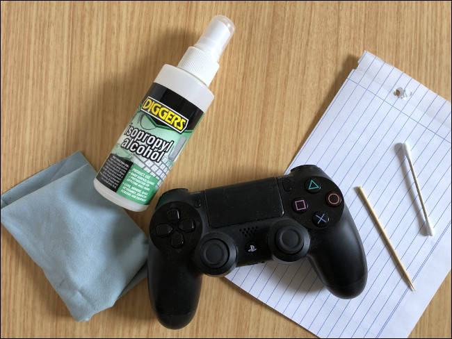 Um DualShock 4 Controller colocado em cima de um pedaço de papel próximo a um palito, um cotonete, um frasco spray de álcool isopropílico e um pano. 