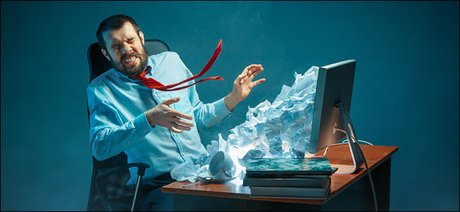 Jovem estressado bonito empresário trabalhando na mesa em um escritório moderno, gritando para a tela do laptop e zangado com o spam de e-mail.  Colagem com uma montanha de papel amassado.