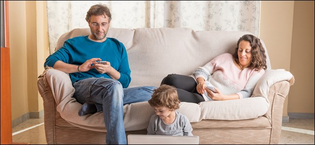 Um homem e uma mulher em um sofá olhando para seus telefones, enquanto uma criança está sentada no chão ao lado deles com um laptop aberto.