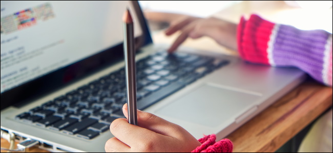 As mãos de uma criança digitando em um MacBook e segurando um lápis.