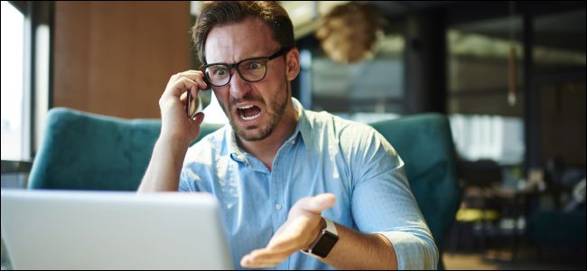 Homem chateado no celular, gesticulando para um laptop.