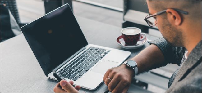 Um homem em uma mesa em um café olhando para o relógio com o laptop aberto na frente dele.