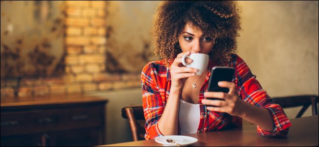 Uma mulher sentada à mesa, olhando para um smartphone e bebendo café.
