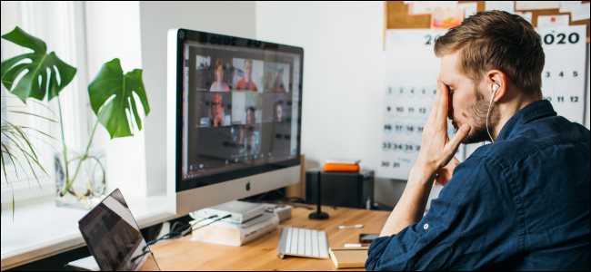 Um homem com a mão no rosto na frente de um desktop e laptop Mac durante uma reunião da Zoom.