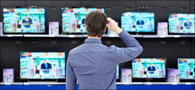 Um homem coçando a cabeça enquanto olha para a tela de TVs em uma loja.