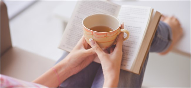 As mãos de uma mulher segurando uma caneca de chá sobre um livro aberto.