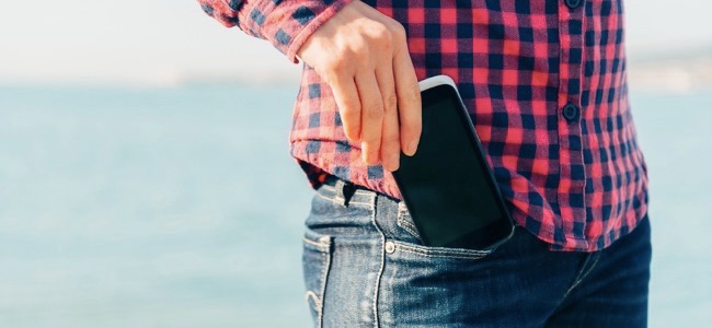 Mulher tira o celular do bolso da calça jeans na praia perto do mar para fazer autorretrato ou para fotografar o mar