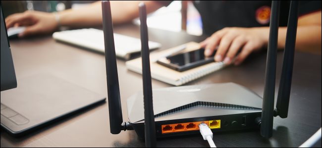 Um roteador sem fio em uma mesa com um homem usando um computador Mac e um iPhone em segundo plano.,