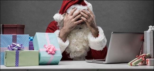 Papai Noel sentado na frente de seu laptop e uma pilha de presentes, cobrindo o rosto com as mãos. 