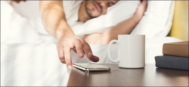 Um homem sonolento na cama, pegando um smartphone na mesa de cabeceira.