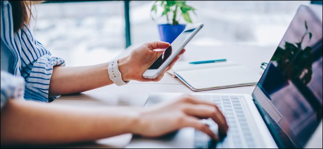 A mão de uma mulher segurando um smartphone enquanto digita no teclado de um MacBook com a outra.