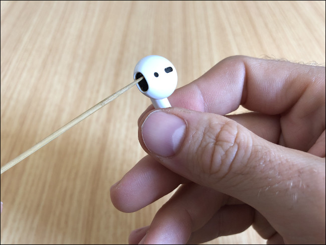 A mão de um homem segurando um AirPod e limpando o alto-falante com o palito.