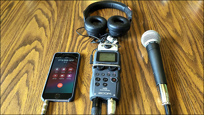 Um iPhone, microfone Shure SM58 e fones de ouvido, todos conectados a um gravador H5 Zoom, sobre uma mesa.