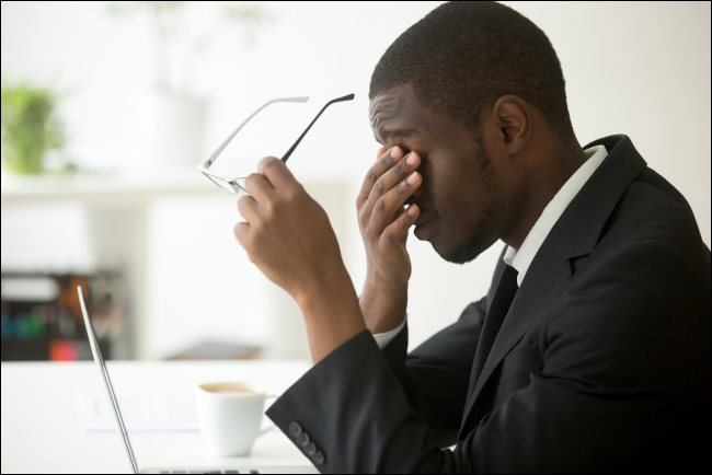 Um homem sentado na frente de seu laptop, segurando os óculos e esfregando os olhos.