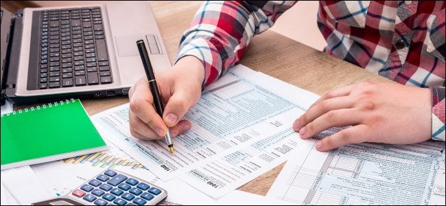 Uma pessoa que preenche 1.040 formulários fiscais em papel perto de um laptop.