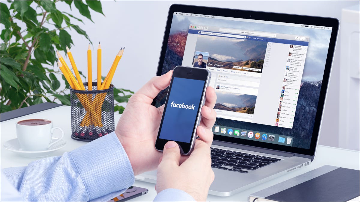 Mãos segurando um smartphone na frente do laptop com o Facebook aberto