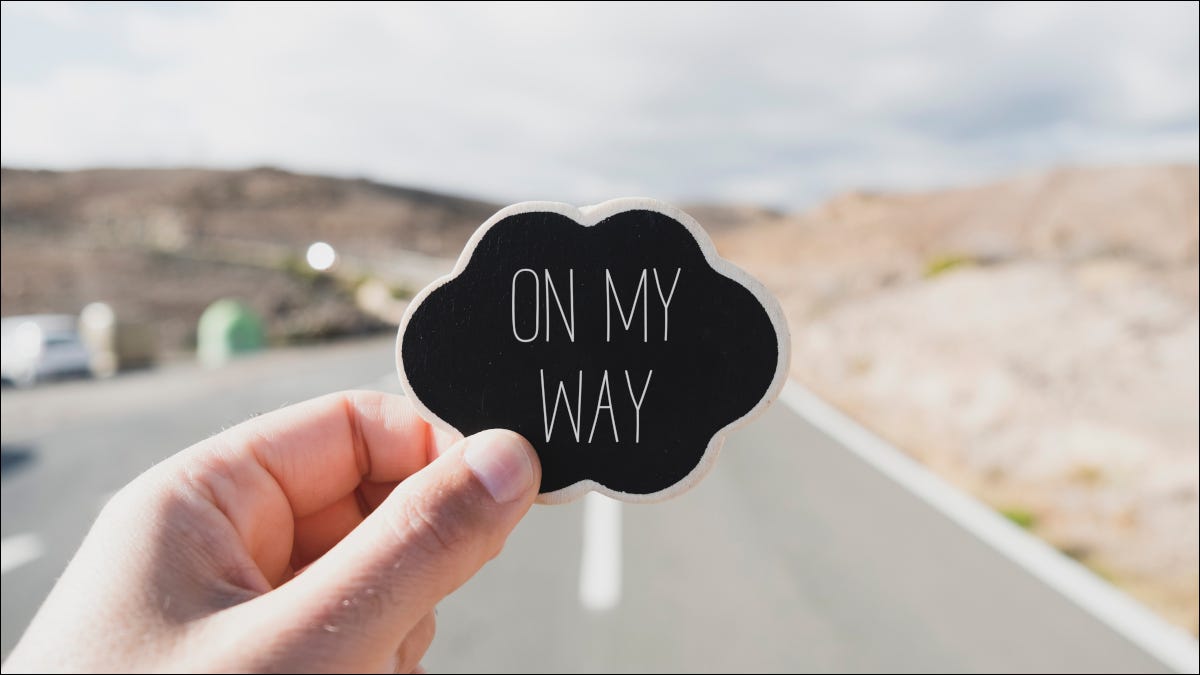 Mão segurando uma pequena placa dizendo "On My Way" na frente de uma estrada.