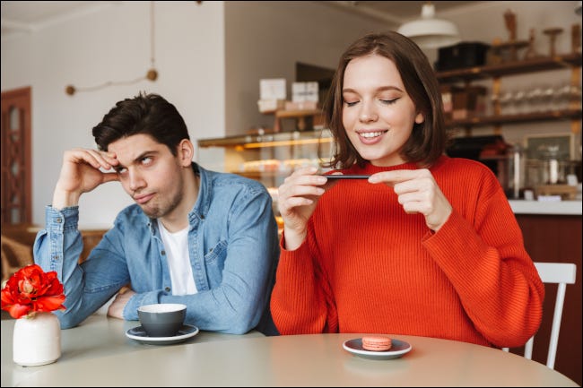 Homem parecendo exasperado ao lado de uma mulher fotografando comida