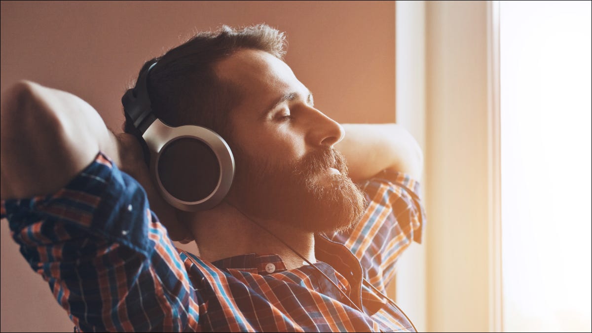 Homem barbudo com as mãos atrás da cabeça, olhos fechados e fones de ouvido, ouvindo áudio.