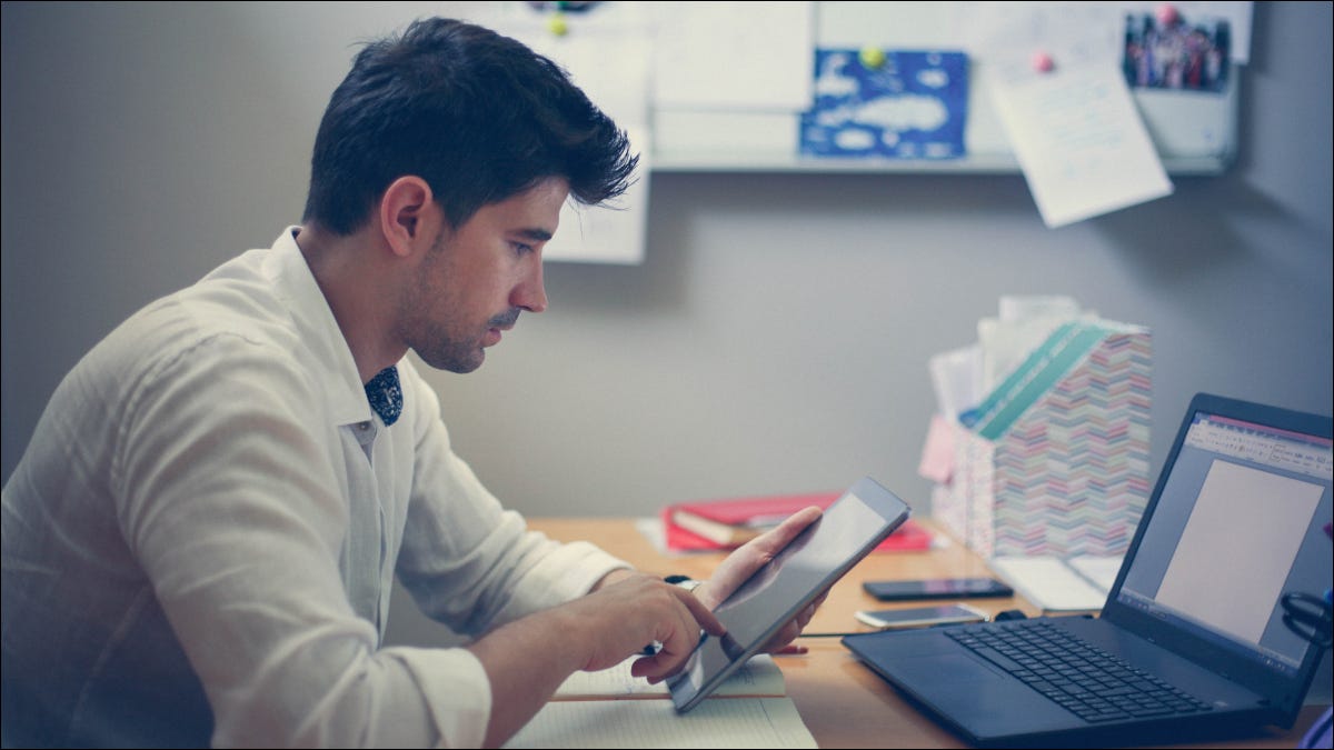 Homem usando um tablet com um laptop na frente dele.