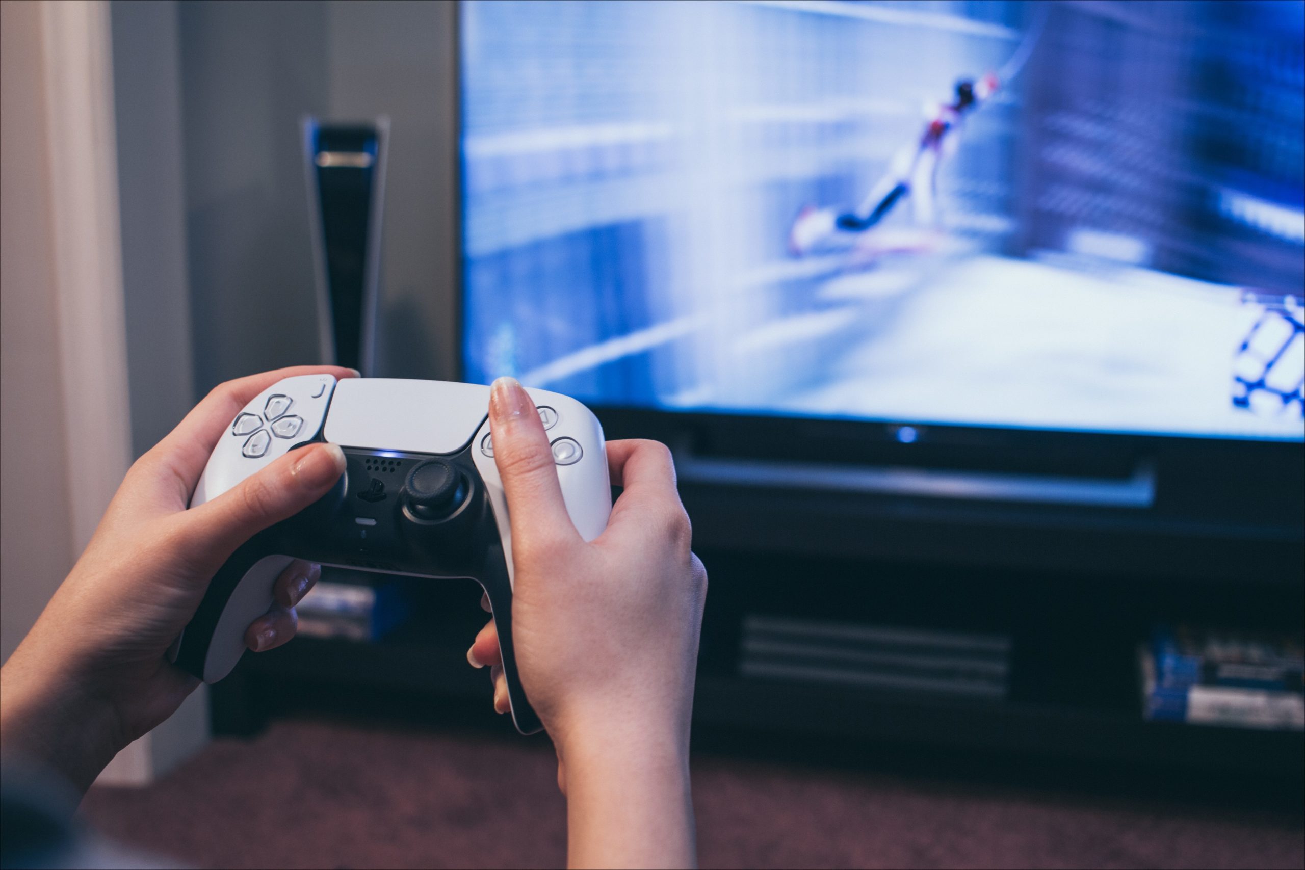 Mãos segurando um controle do PlayStation 5 na frente de uma TV.