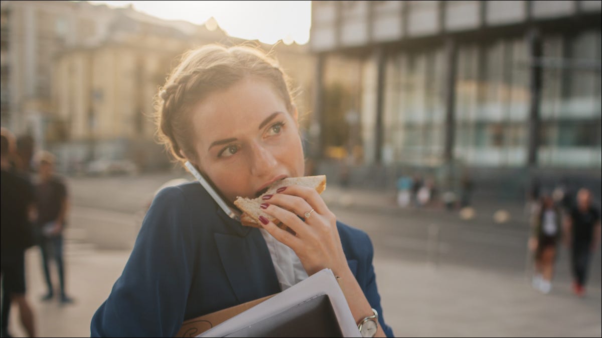 Mulher ocupada comendo um sanduíche e falando ao celular ao mesmo tempo.