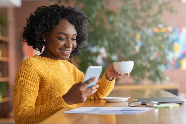 Mulher tomando café e sorrindo enquanto olha para o smartphone