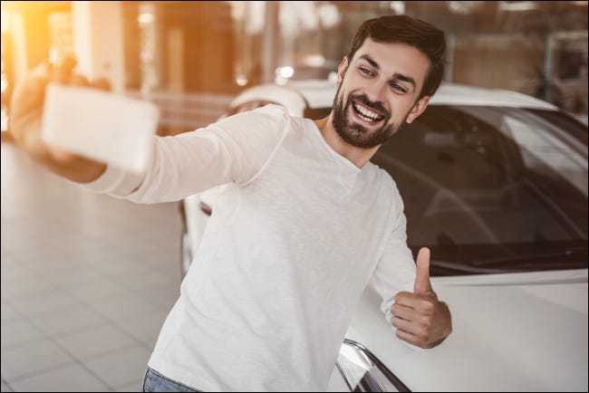 Jovem tirando uma selfie e dando um polegar para cima na frente de um carro.