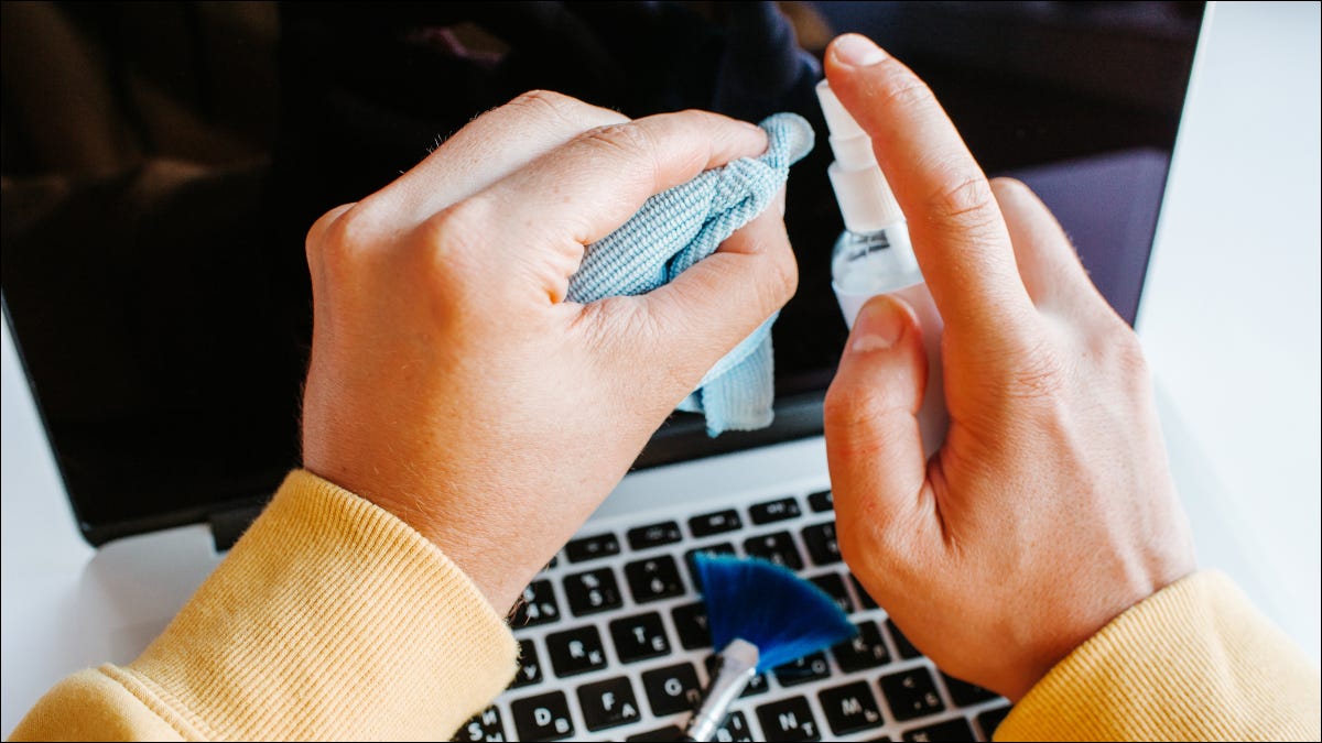 Mãos segurando um pano de microfibra e spray de limpeza sobre um laptop aberto com uma escova sobre o teclado.