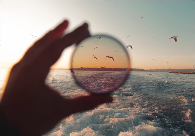 Mão segurando um filtro de lente para demonstrar o efeito de polarização em uma cena de praia.