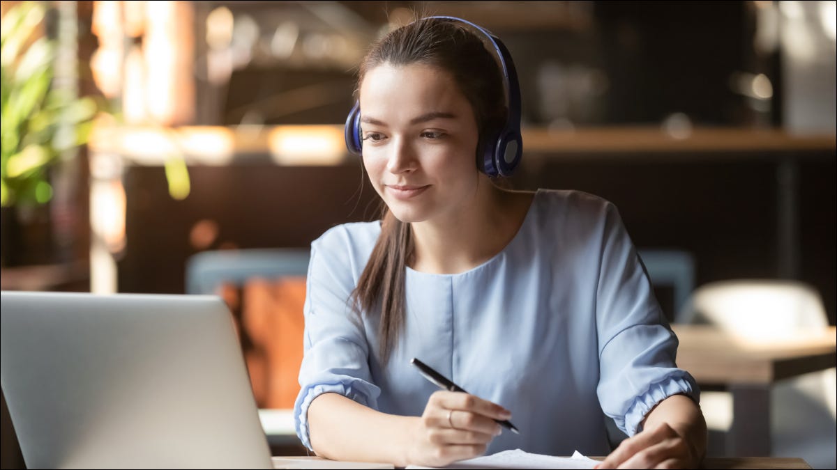 Jovem mulher usando fones de ouvido enquanto toma notas na frente de um laptop,