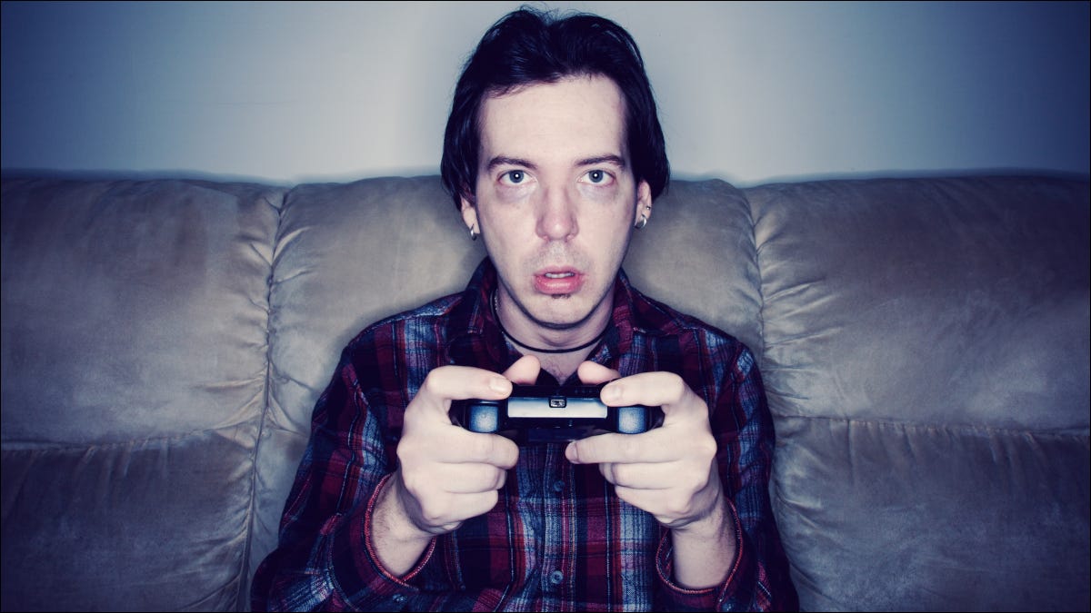 Homem sentado em um sofá e jogando videogame com uma expressão exausta ou atordoada.