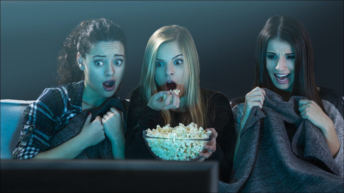 Três jovens assistindo a um filme e comendo pipoca com expressões assustadas.