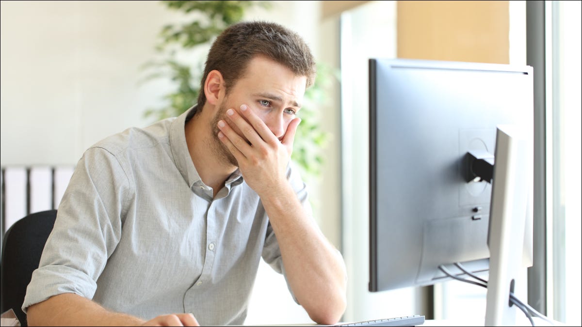 Homem olhando para o computador com expressão chocada, cobrindo a boca.