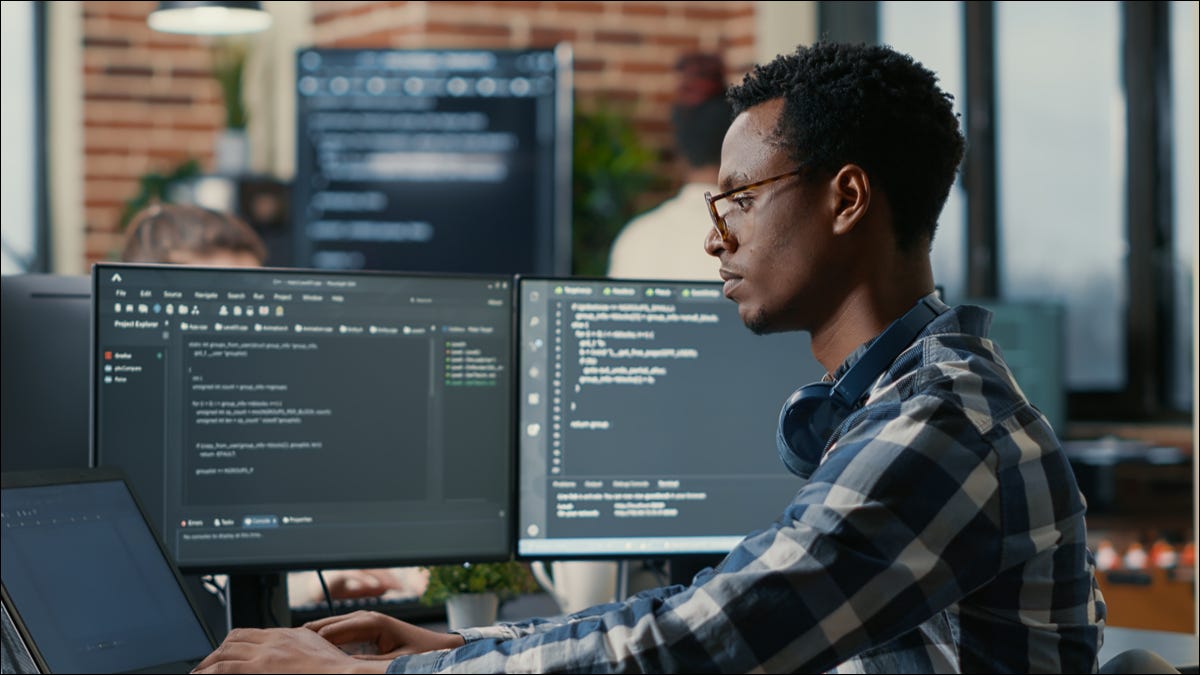 Você homem usando um laptop ao lado de vários monitores de computador com código em exibição.