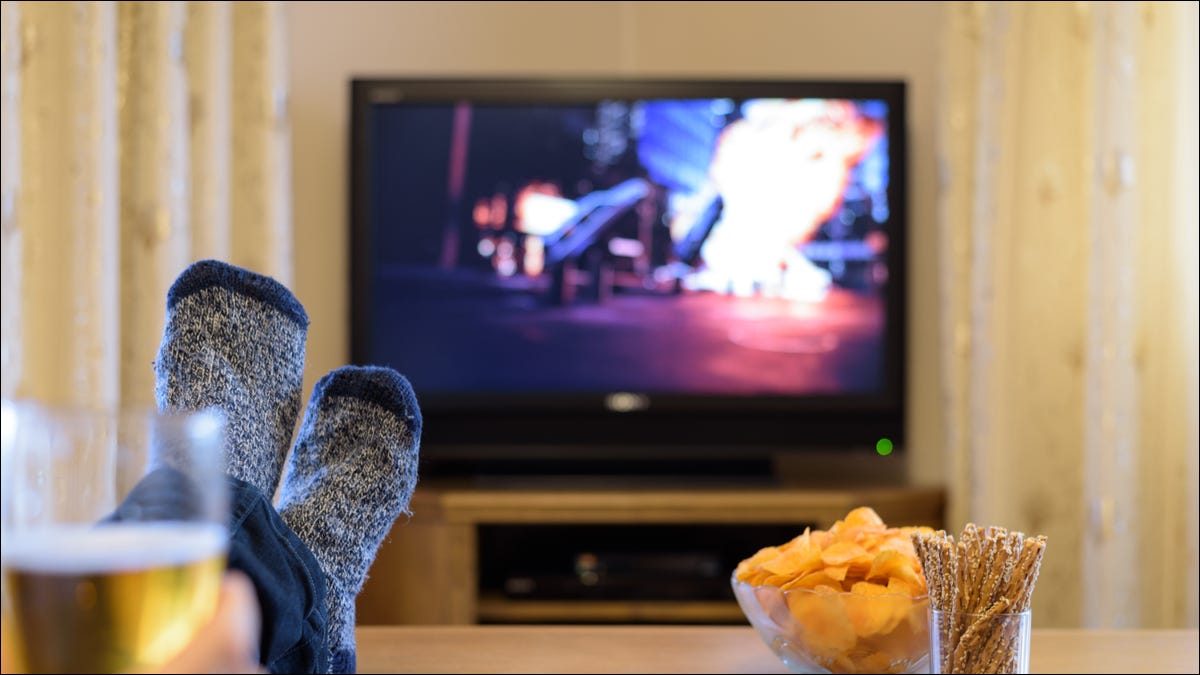 Os pés de uma pessoa e lanches em uma mesa de centro em frente a uma TV com um filme de ação em exibição.