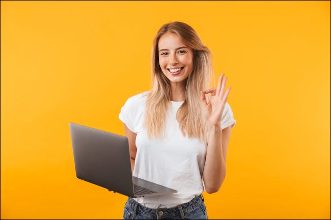 Uma jovem sorrindo e fazendo um gesto de "ok" com a mão enquanto segura um laptop.