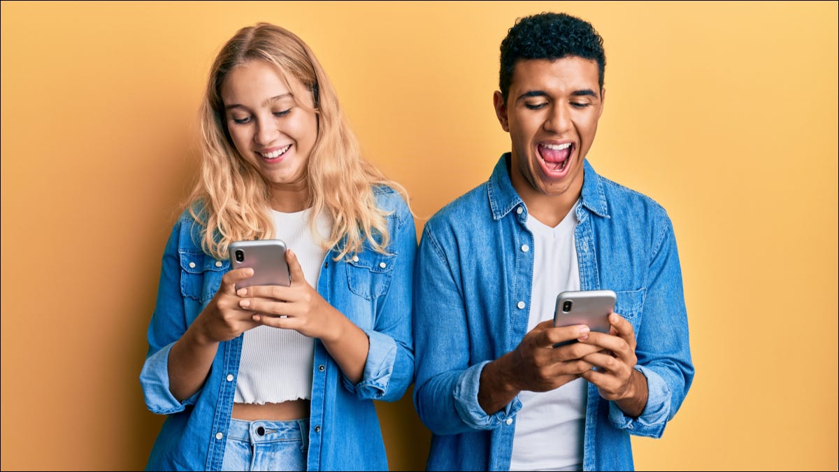 Um jovem casal ao lado um do outro enquanto sorrindo e usando seus smartphones.