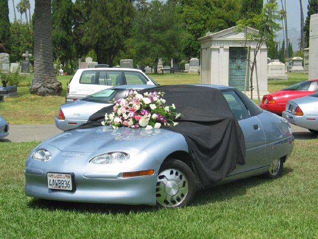 Foto de vários EV1 estacionados em um cemitério, com flores e uma cortina cobrindo um dos carros