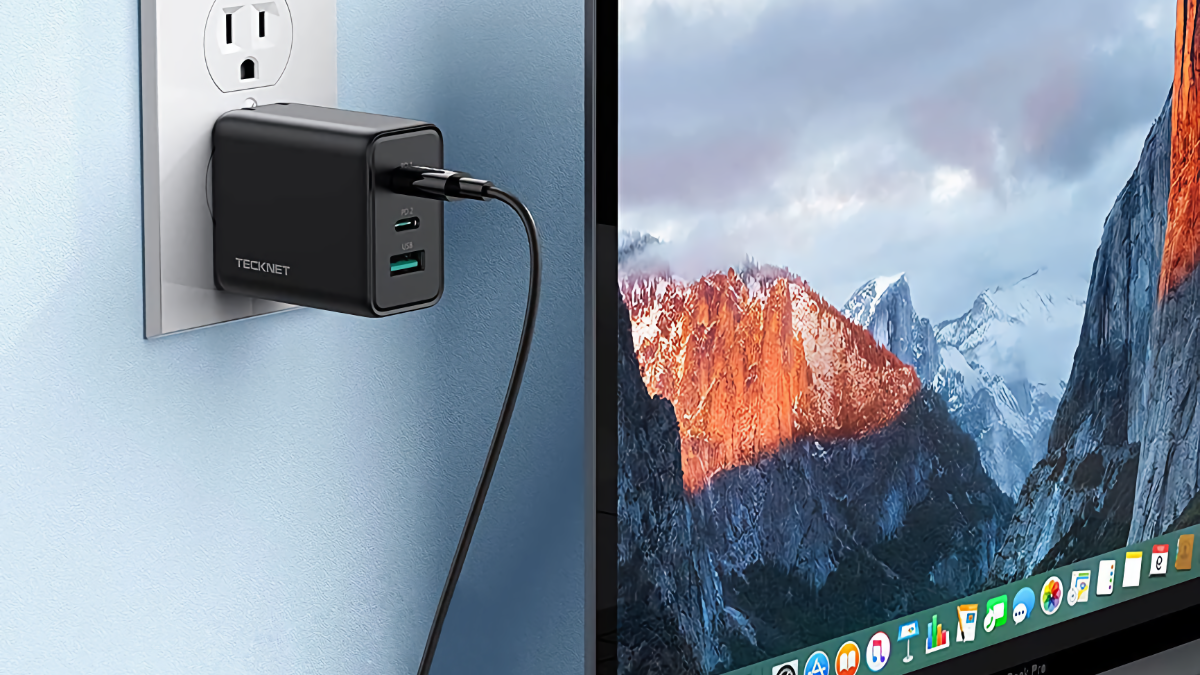 Carregador USB-C TECKNET 65W conectado a uma tomada ao lado de um MacBook